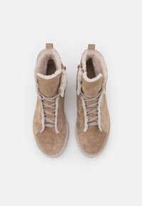 Bottines en suède beige avec un laçage à l'avant, doublure intérieure en peluche et fermeture éclair sur le côté. Surface légèrement texturée avec des détails cousus autour de la semelle.