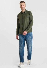 Man staat, draagt een olijfgroene overhemd met knopen, blauwe cargobroeken met patchzakken en witte sportschoenen.