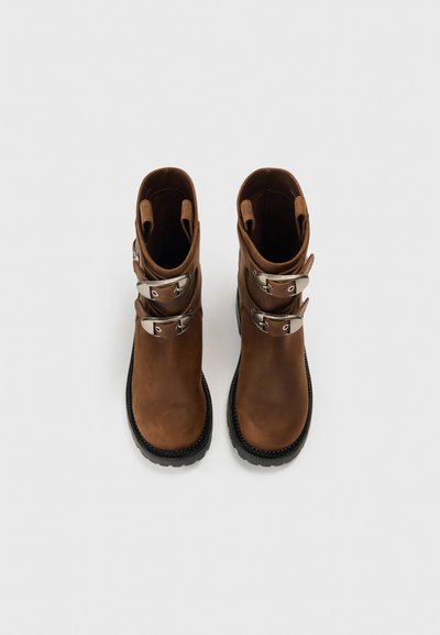 Hnědé kožené kotníkové boty se dvěma stříbrnými přezkami, strukturovanou gumovou podrážkou a zaoblenou špičkou. Hladký svršek a kontrastní černá podrážka.