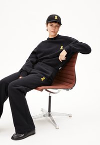 Zwart sweatshirt en broek set met gele lint logo detail. Model zittend in een bruine gestoffeerde stoel, met een bijpassende zwarte pet.