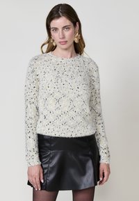 Weißer strukturierter Pullover mit schwarzen und goldenen Sprenkeln, runder Ausschnitt, lange Arme, kombiniert mit einem schwarzen Lederminirock.
