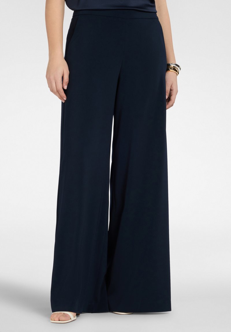 Donna che indossa pantaloni blu navy a vita alta e gamba larga, abbinati a sandali bianchi open-toe e braccialetti dorati su un polso.