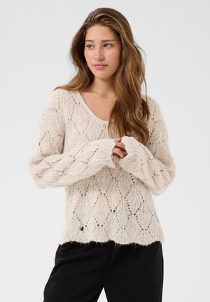 Jeune femme aux longs cheveux bruns porte un pull crème tricoté ample et un pantalon noir, regardant légèrement vers sa gauche avec les mains jointes.