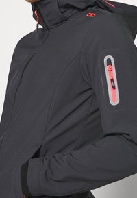 Veste softshell noire avec une poche zippée sur la manche gauche, présentant des accents rouges sur la fermeture éclair et la capuche. Finition mate, design ajusté.