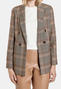 Blazer croisé à carreaux marron, avec des boutons marron, une coupe ajustée et des épaules structurées. Associé à un pantalon en simili cuir marron clair.