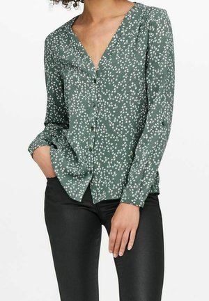 Femme portant un chemisier vert à manches longues avec des motifs floraux blancs et un pantalon slim noir, une main dans la poche.