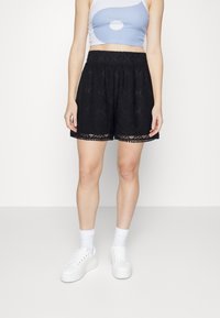 Pantalones cortos de encaje negro con una cinturilla elástica, con un dobladillo festoneado y un intrincado patrón de diamantes. Llevados con zapatillas blancas y calcetines.