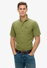 Camisa de botón de manga corta, color oliva, con un patrón texturizado, dos bolsillos en el pecho y botones marrones. Combinada con un cinturón marrón y vaqueros.