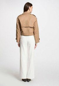 Chaqueta beige recortada con una cintura ajustable, mangas abotonadas y un amplio cuello, combinada con pantalones blancos de cintura alta y botas de tacón marrones.