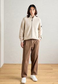 Pull en maille beige zippé avec un design côtelé, associé à un pantalon large marron et des baskets blanches sur un sol en bois.