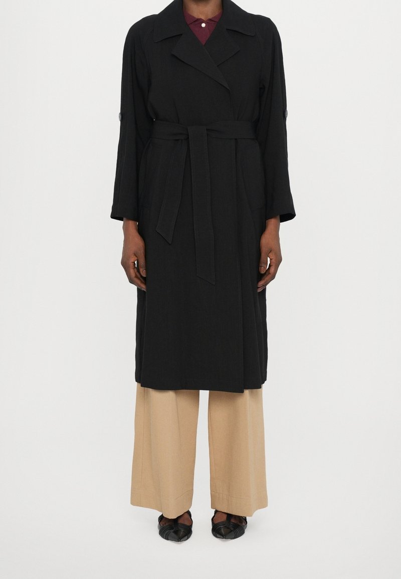 Personne portant un long manteau noir ceinturé, un pantalon beige à jambes larges et des chaussures noires pointues, debout devant un fond blanc uni.