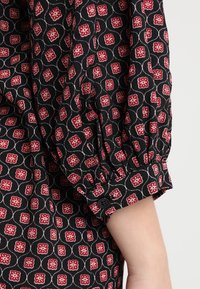 Black fabric with a red and white floral geometric pattern, featuring a gathered elastic cuff with a button detail.