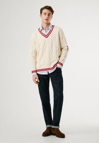 Jovem vestido com um suéter creme de malha com riscas vermelhas, camisa azul clara, jeans escuros e botas de camurça castanhas, de pé com a mão no bolso.