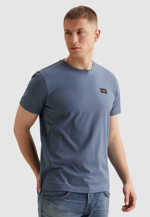 Hombre vistiendo una camiseta azul de manga corta lisa y jeans azules, mirando hacia su izquierda contra un fondo claro y liso.