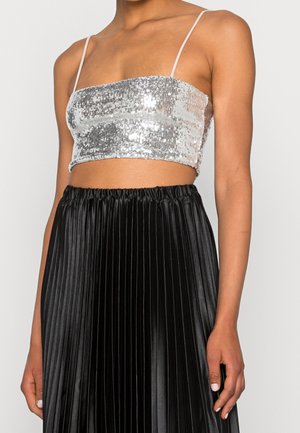 Frau trägt ein silbernes Pailletten-Crop-Top mit dünnen Trägern und einen schwarzen, hoch taillierten, plissierten Rock.