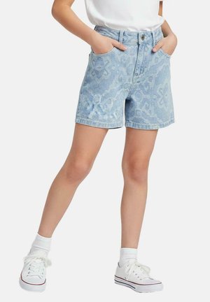 Kind draagt lichtblauwe spijkershorts met patroon, witte sokken en witte sportschoenen, staat met handen in de zakken.