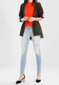 Olive green water-resistant jacket, red T-shirt, light blue ripped skinny jeans, black and white block-heeled shoes, casual outfit.