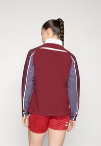 Maroon jacket with striped blue sleeves and white accents, featuring a high collar. Red shorts with logo detail complete the outfit.
