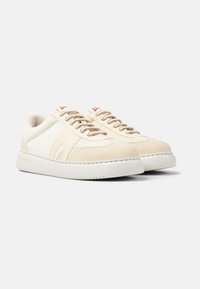 Beige Sneaker mit Wildleder- und Mesh-Einsätzen, runder Zehenpartie, flacher weißer Sohle, beigen Schnürsenkeln und rotem Logo-Akzent auf der Zunge.