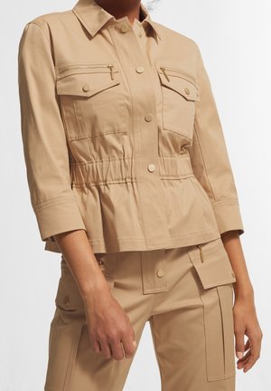 Veste utilitaire beige avec boutons-pression, poches poitrine zippées, taille élastique, associée à un pantalon cargo assorti avec poches à rabat.