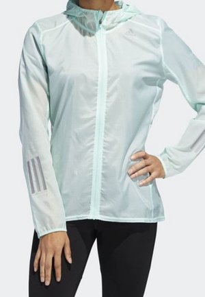 Osoba ubrana w jasnoniebieską kurtkę z kapturem na zamek błyskawiczny z trzema odblaskowymi paskami na lewym rękawie oraz czarne legginsy, stojąca z ręką na biodrze.