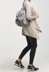 Sac à dos en tissu gris avec des sangles en cuir brun et des éléments en métal, équipé d'un rabat avant et de deux poches latérales. Porté sur une épaule.