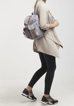 Sac à dos en tissu gris avec des sangles en cuir brun et des éléments en métal, équipé d'un rabat avant et de deux poches latérales. Porté sur une épaule.
