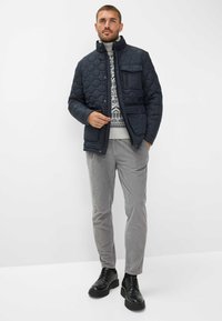 Giacca trapuntata blu navy con colletto alto, tasca con zip, indossata sopra un maglione grigio a fantasia, abbinata a pantaloni grigi chiari e scarpe nere.