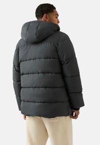 Pufferjack in donkergrijs met horizontale doorgestikte textuur, een capuchon en elastische manchet, gedragen over crèmekleurige broek.