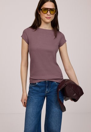 Donna con occhiali dalle lenti gialle, top mauve a maniche corte, jeans blu, che tiene in mano destra una giacca in pelle color bordeaux scuro piegata.