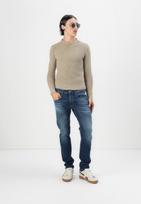 Maglione beige a coste, jeans blu e sneakers bianche con accenti in beige. L'outfit presenta una vestibilità comoda e uno stile casual.