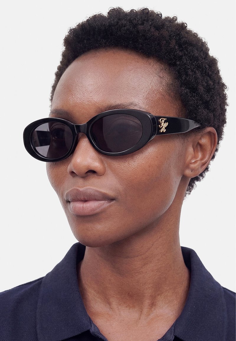 Personne aux cheveux courts et bouclés portant des lunettes de soleil ovales noires avec un logo doré sur le côté et une chemise sombre à col.
