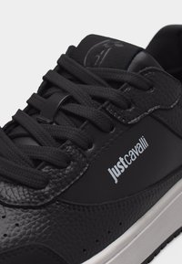 Črni teksturirani usnjeni športni copat s črnimi vezalkami, belim logotipom Just Cavalli na strani in belim podplatom na enostavni svetli podlagi.