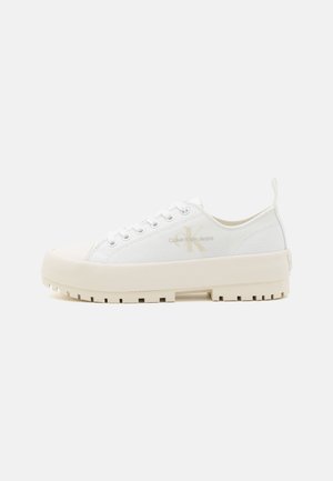 Zapatillas de lona blancas con una suela de goma cremosa gruesa y un acabado texturizado. Presentan cordones y un logo en el lateral.