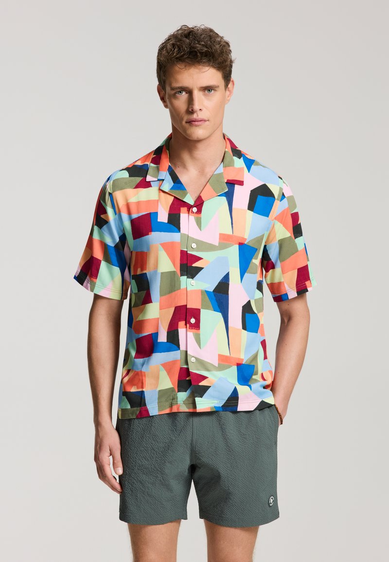 Mann trägt ein buntes, kurzärmeliges Hemd mit geometrischem Muster und strukturierte dunkelgrüne Shorts, steht mit den Händen in den Taschen vor einfachem Hintergrund.