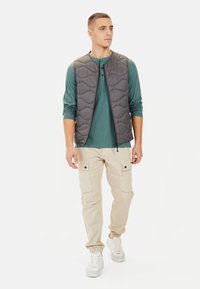 Herre grå quiltet puffer vest over en grøn langærmet skjorte, parret med beige cargo bukser med lommer og elastiske manchetter, iført hvide sneakers.