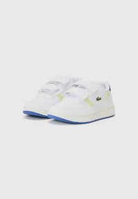 Hvite sneakers med borrelåsremmer, grønn akseentstripe, teksturert gummisåle og blå detaljer på sålen. Lacoste-logo synlig.