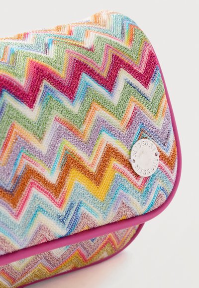 Έγχρωμο πλεκτό μοτίβο ζιγκ-ζαγκ σε ύφασμα με κυκλικό λευκό μεταλλικό ταμπελάκι με χαραγμένο το "MISSONI", περιτριγυρισμένο από ροζ περίγραμμα.