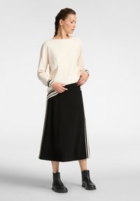 Cremefarbenes Sweatshirt mit schwarzen Streifen an den Bündchen, kombiniert mit einem schwarzen Rock mit Seitenstreifen. Das Model trägt schwarze Stiefeletten.