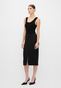 Débardeur noir à côtes avec bretelles larges associé à une jupe crayon noire avec une fente sur le côté. Modèle portant des chaussures à talons hauts noires.