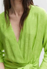 Grüne Wickelbluse mit tiefem V-Ausschnitt, die mit strukturierten floralen Mustern, langen Ärmeln und einem gebundenen Taillendetail versehen ist. Sanfter Stofffinish.