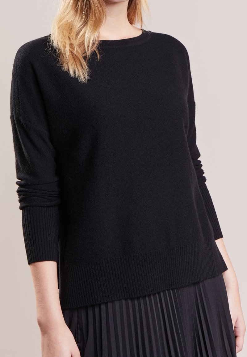Femme portant un pull en maille noir à manches longues avec poignets et ourlet côtelés, associé à une jupe plissée noire sur un fond neutre.
