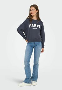 Sweatshirt navy con le scritte "PARIS" e "FRANCE" in lettere bianche, maniche lunghe, abbinato a jeans a zampa d'elefante blu chiaro e sneakers bianche.