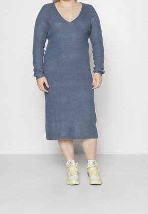 Femme portant une robe midi en tricot bleu à manches longues avec un col en V et des baskets montantes jaune clair, debout devant un fond uni.