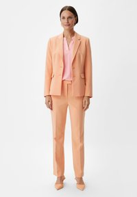 Pfirsichfarbener Anzug aus glattem Stoff. Blazer mit zwei Knöpfen und schmal geschnittene Hose. Hellrosa Bluse darunter. Spitz zulaufende pfirsichfarbene High Heels.