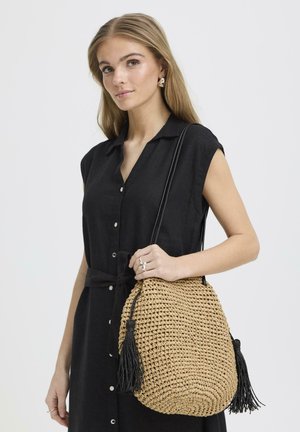 Joven mujer con cabello largo y rubio que lleva un vestido negro sin mangas con botones, sosteniendo un bolso tejido color canela con borlas negras.