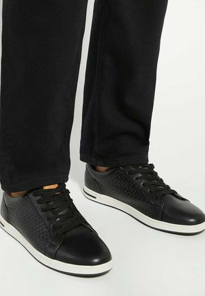 Baskets noires avec une tige en cuir synthétique texturé et en cuir lisse, dotées d'une semelle en caoutchouc blanche et de lacets noirs, conçues pour un usage décontracté.