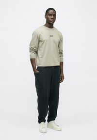 Μπεζ φούτερ με λογότυπο, μακριά μανίκια και μανσέτες, συνδυασμένο με σκούρο πράσινο παντελόνι με πλαϊνές τσέπες. Το μοντέλο φορεί λευκά αθλητικά παπούτσια.