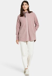 Flauschige, hellrosa Fleecejacke mit Reißverschluss, hohem Kragen, langen Ärmeln und kontrastierendem Reißverschluss; kombiniert mit weißen Hosen und hellen Sneakers.