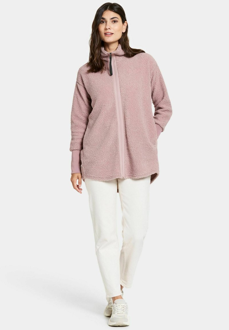 Flauschige, hellrosa Fleecejacke mit Reißverschluss, hohem Kragen, langen Ärmeln und kontrastierendem Reißverschluss; kombiniert mit weißen Hosen und hellen Sneakers.